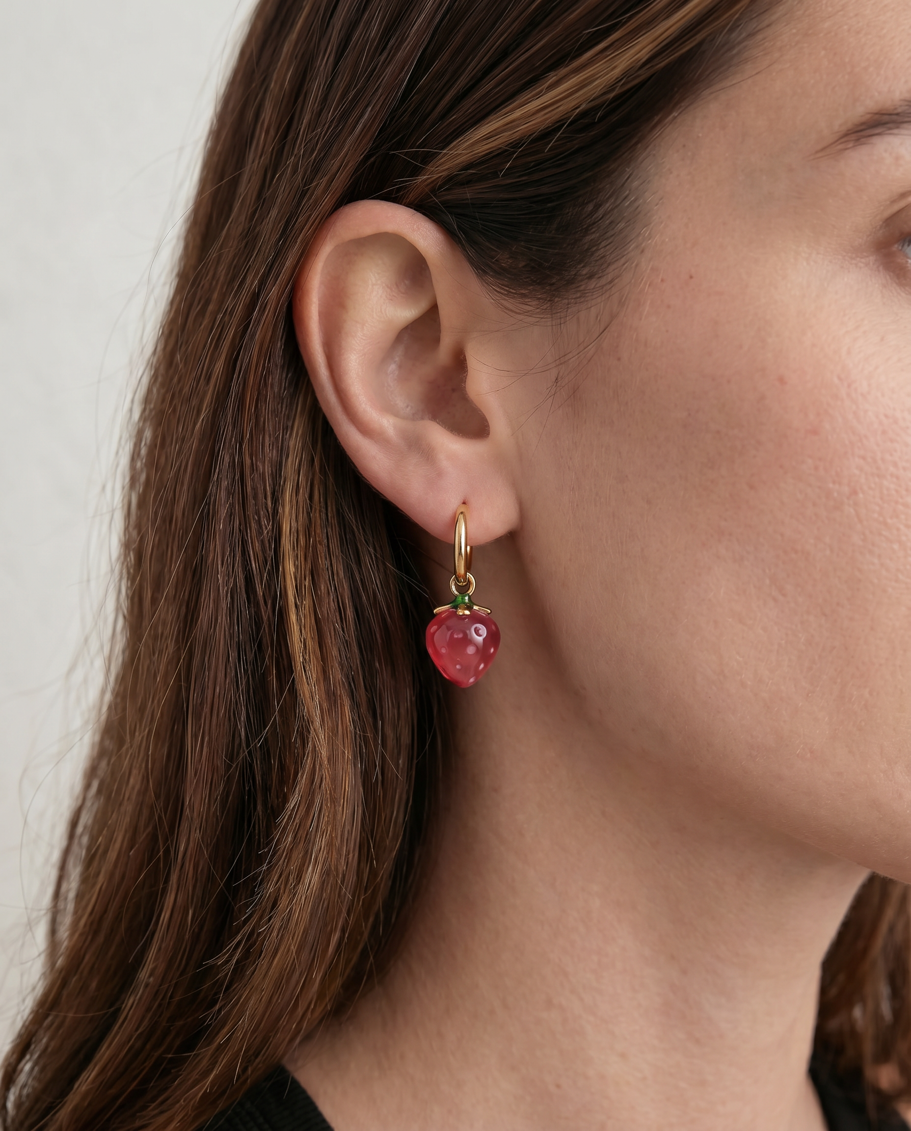 Close-up of a person wearing a gold earring with a red strawberry gemstone against a neutral background