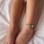 A close-up of a child's legs resting on white bedding, wearing a delicate bracelet with a light green jade disc and small pearl-like beads.