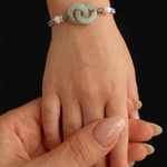 A close-up of a delicate beaded bracelet with a central pale green jade-like disc and white pearls, worn on a person's wrist while another hand gently holds it against a solid black background.