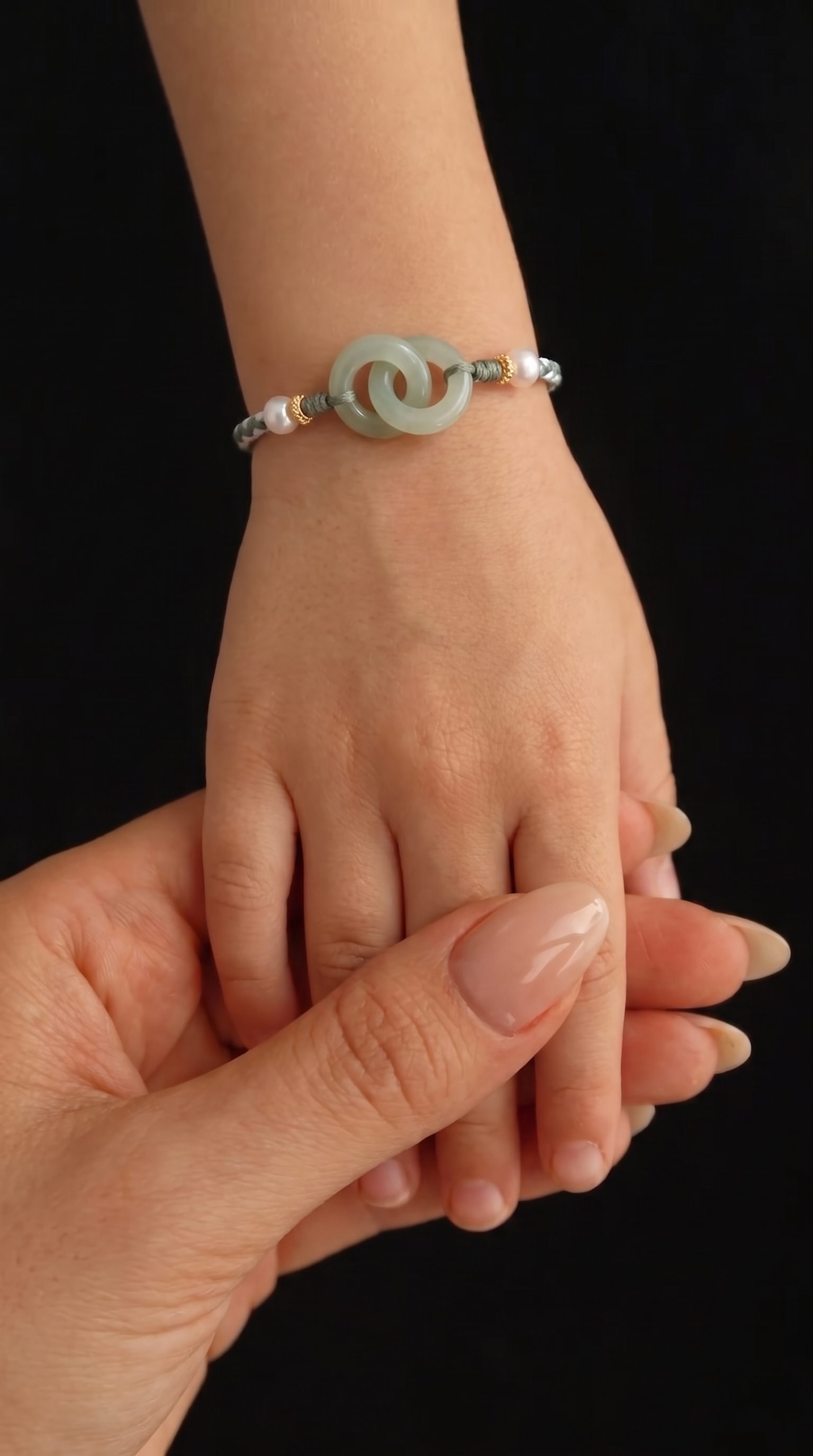 A close-up of a delicate beaded bracelet with a central pale green jade-like disc and white pearls, worn on a person's wrist while another hand gently holds it against a solid black background.