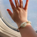 A child's hand resting on an airplane window, wearing a beaded bracelet with a central white jade-like ring and gold-toned beads.