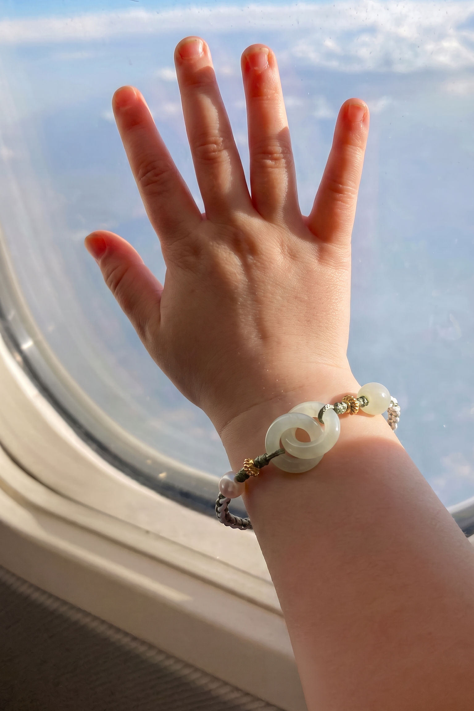 A child's hand resting on an airplane window, wearing a beaded bracelet with a central white jade-like ring and gold-toned beads.