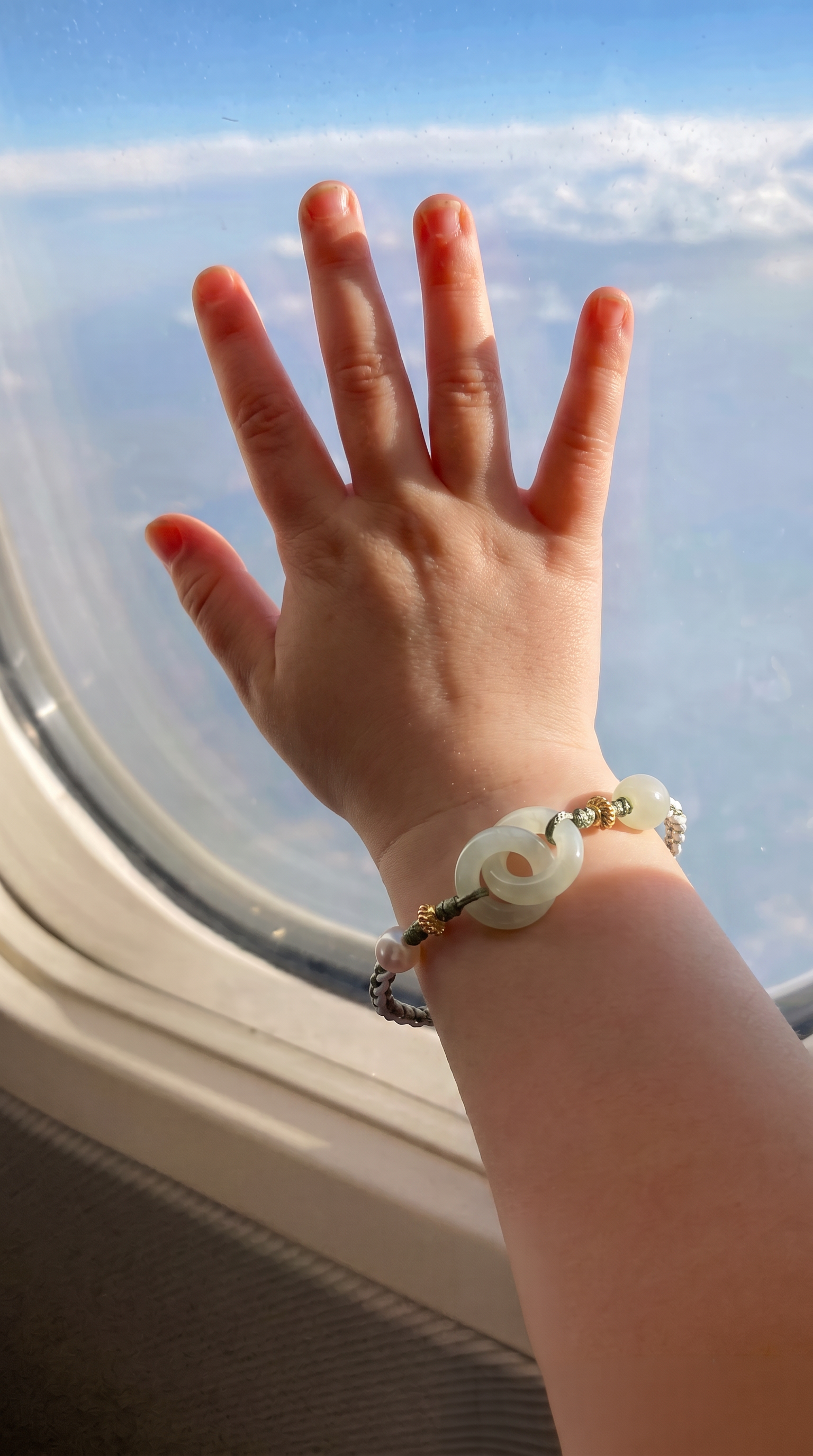 A child's hand resting on an airplane window, wearing a beaded bracelet with a central white jade-like ring and gold-toned beads.