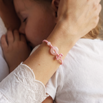 A close-up lifestyle shot of a young child hugging an adult, with the adult's hand gently holding the child's head. The adult is wearing a delicate pink beaded bracelet with a central pink stone and a small bow detail on the wrist.