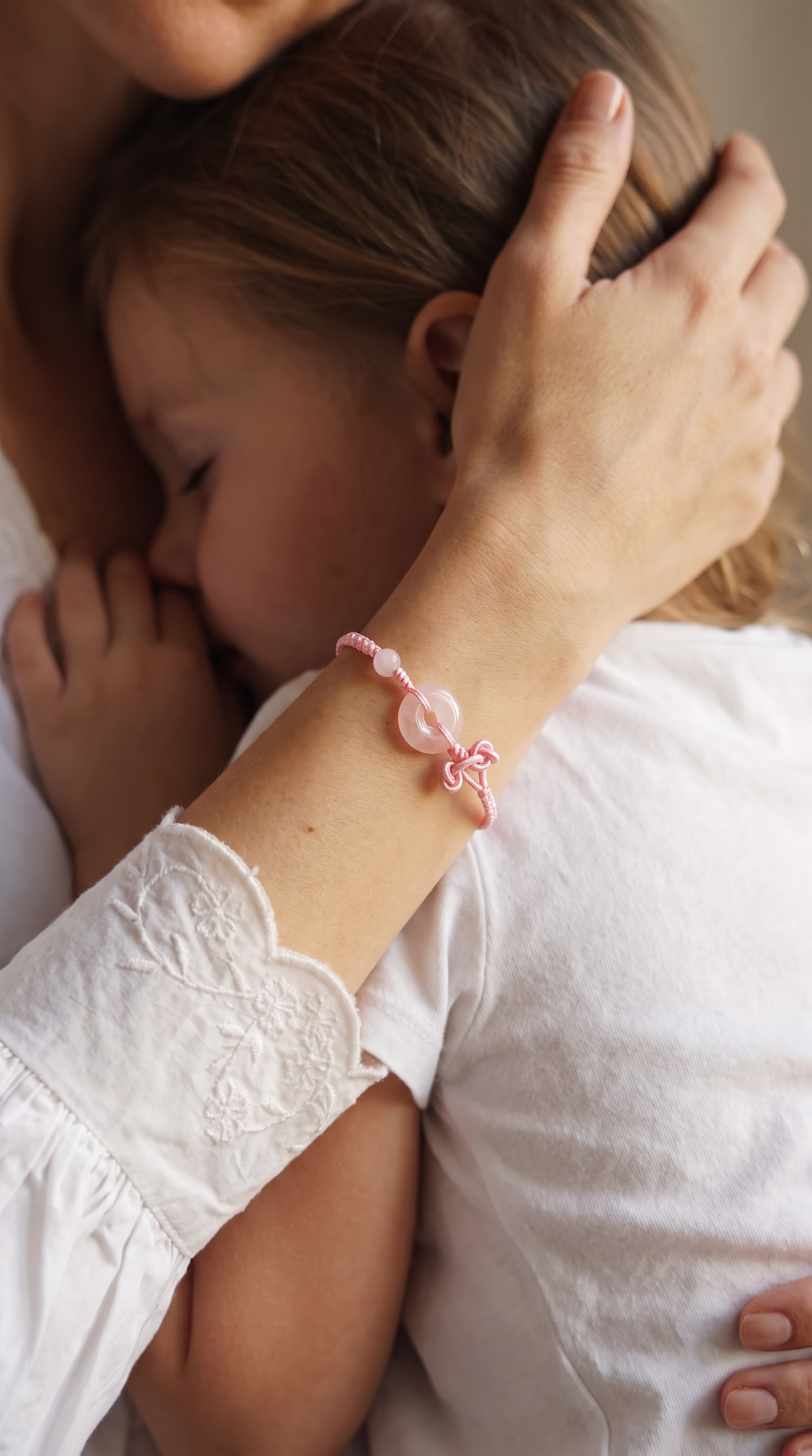 A close-up lifestyle shot of a young child hugging an adult, with the adult's hand gently holding the child's head. The adult is wearing a delicate pink beaded bracelet with a central pink stone and a small bow detail on the wrist.