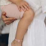 A close-up lifestyle shot of a mother in a white linen dress holding her child, who is wearing light pink shorts and a pink beaded anklet.