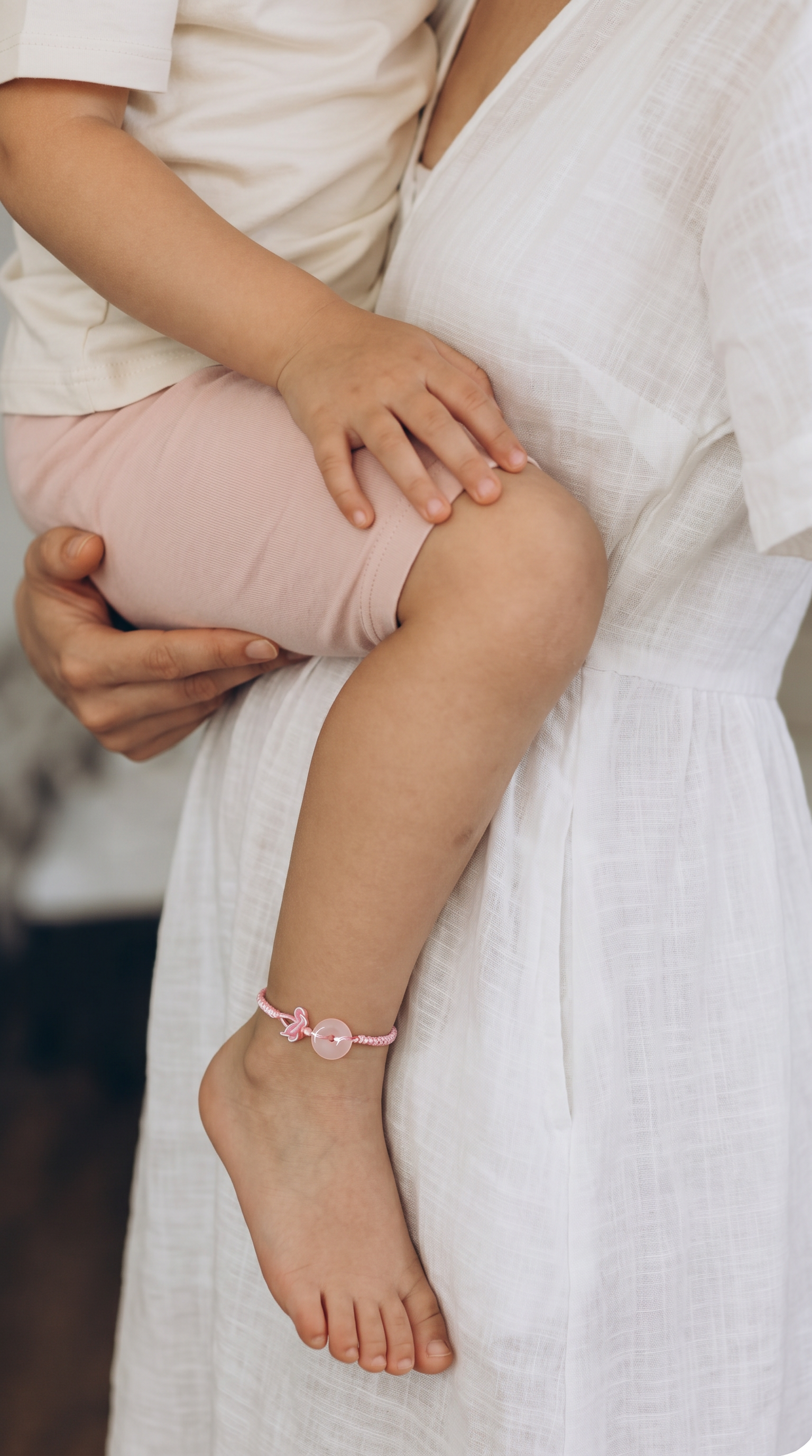 A close-up lifestyle shot of a mother in a white linen dress holding her child, who is wearing light pink shorts and a pink beaded anklet.