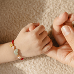 A close-up of a child's hand holding an adult's hand, with the child's wrist wearing a red cord bracelet decorated with a light green jade-like bead, a small gold charm, and a white pearl.