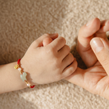 A close-up of a child's hand holding an adult's hand, with the child's wrist wearing a red cord bracelet decorated with a light green jade-like bead, a small gold charm, and a white pearl.