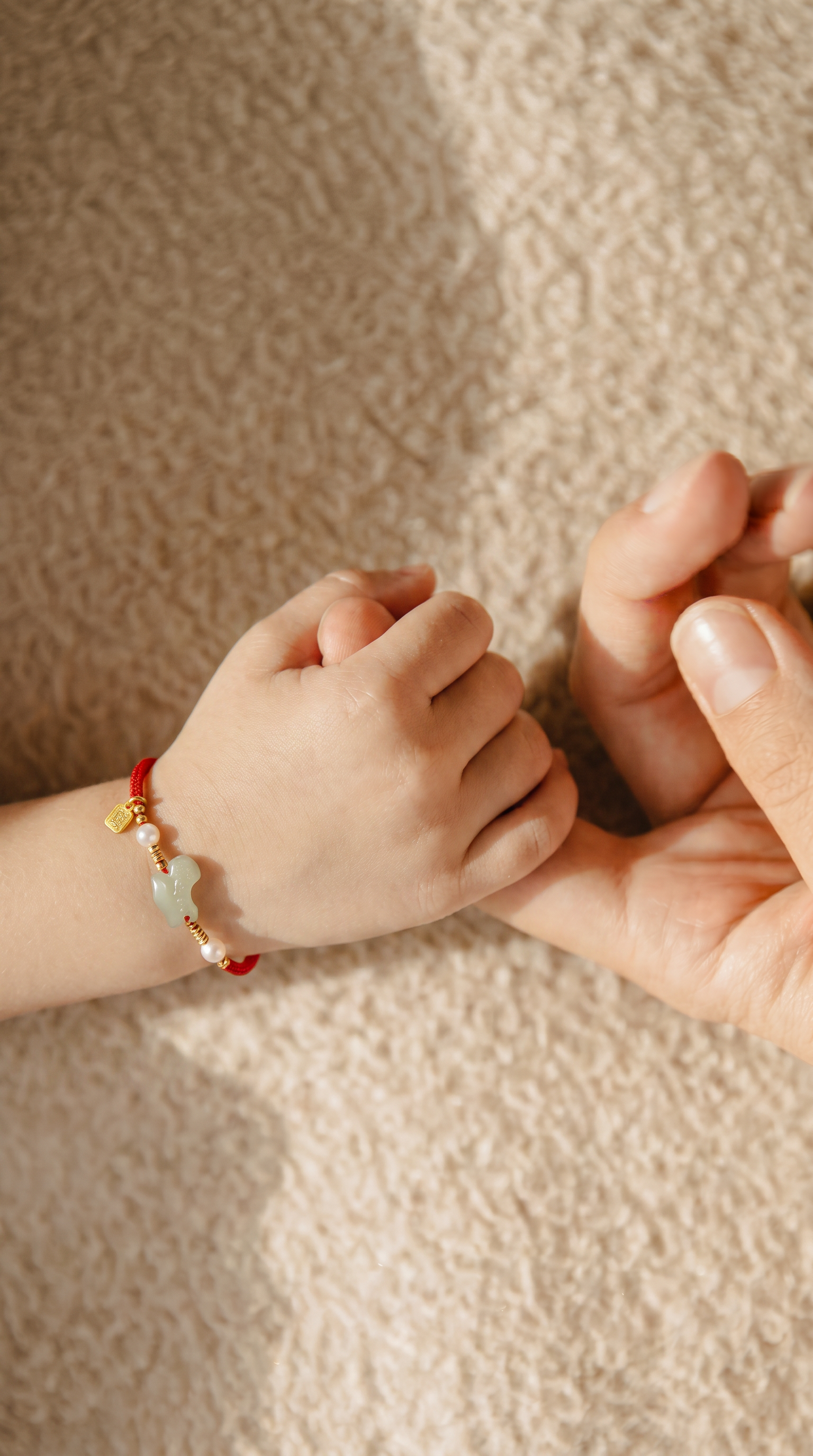A close-up of a child's hand holding an adult's hand, with the child's wrist wearing a red cord bracelet decorated with a light green jade-like bead, a small gold charm, and a white pearl.