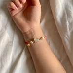 A close-up of a person's wrist wearing a delicate red cord bracelet with gold-toned charms, including a small heart, a pearl-like bead, and a light stone, resting on white fabric.