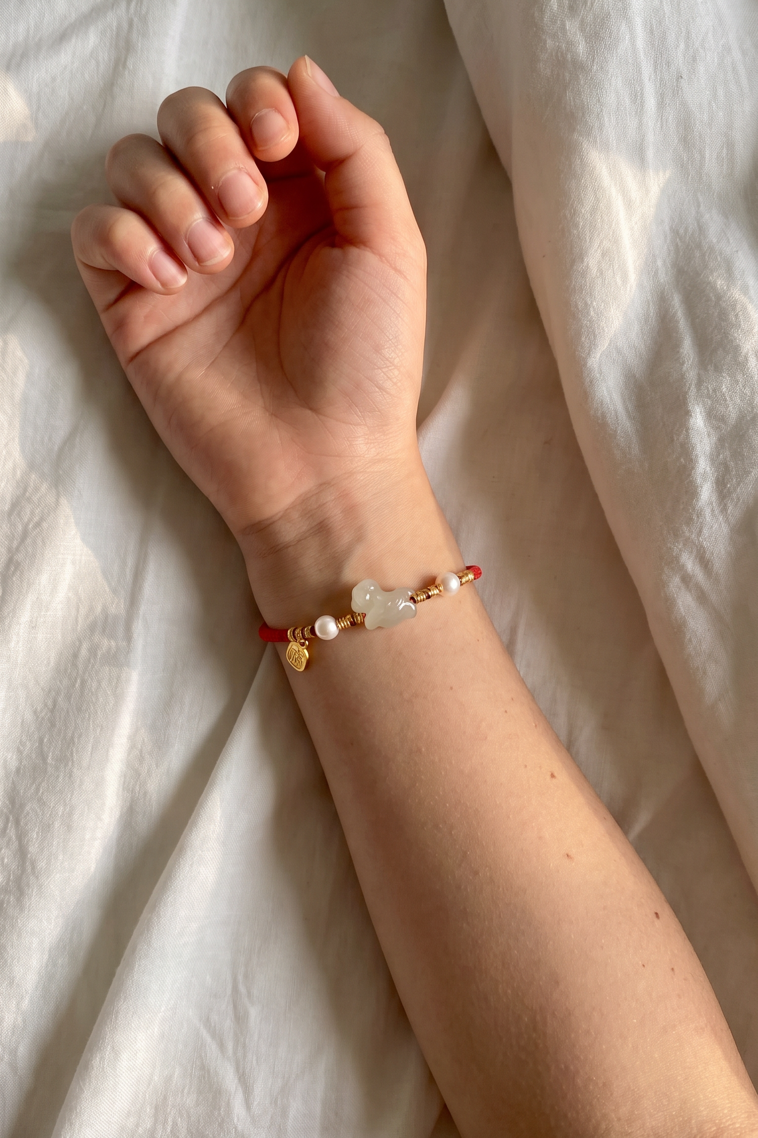A close-up of a person's wrist wearing a delicate red cord bracelet with gold-toned charms, including a small heart, a pearl-like bead, and a light stone, resting on white fabric.