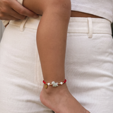 A close-up lifestyle shot of a baby's bare foot and ankle wearing a red cord bracelet with a light green jadeite cloud charm and gold accents, while being held by an adult wearing a white ribbed top and white pants.