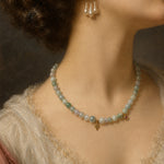 A close-up of a woman's neck and upper chest wearing a delicate beaded necklace with pale green and white pearls, gold-toned accents, and matching pearl drop earrings.