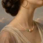 A close-up portrait of a woman wearing a delicate pearl necklace with small green, orange, and white accent beads, along with large gold hoop earrings and a sheer cream dress against a soft painted sky background.