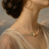A close-up portrait of a woman wearing a delicate pearl necklace with small green, orange, and white accent beads, along with large gold hoop earrings and a sheer cream dress against a soft painted sky background.