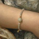 A close-up of a delicate beaded bracelet worn on a person's wrist. The bracelet features pale green and white beads with small gold spacer beads and a small gold charm, set against a painterly gray and white background.